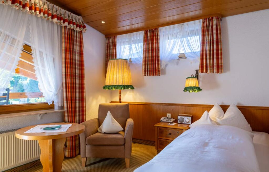 Einzelzimmer mit karierten Vorhängen, Sessel, Bett und Tisch. Rustikal-gemütlich.